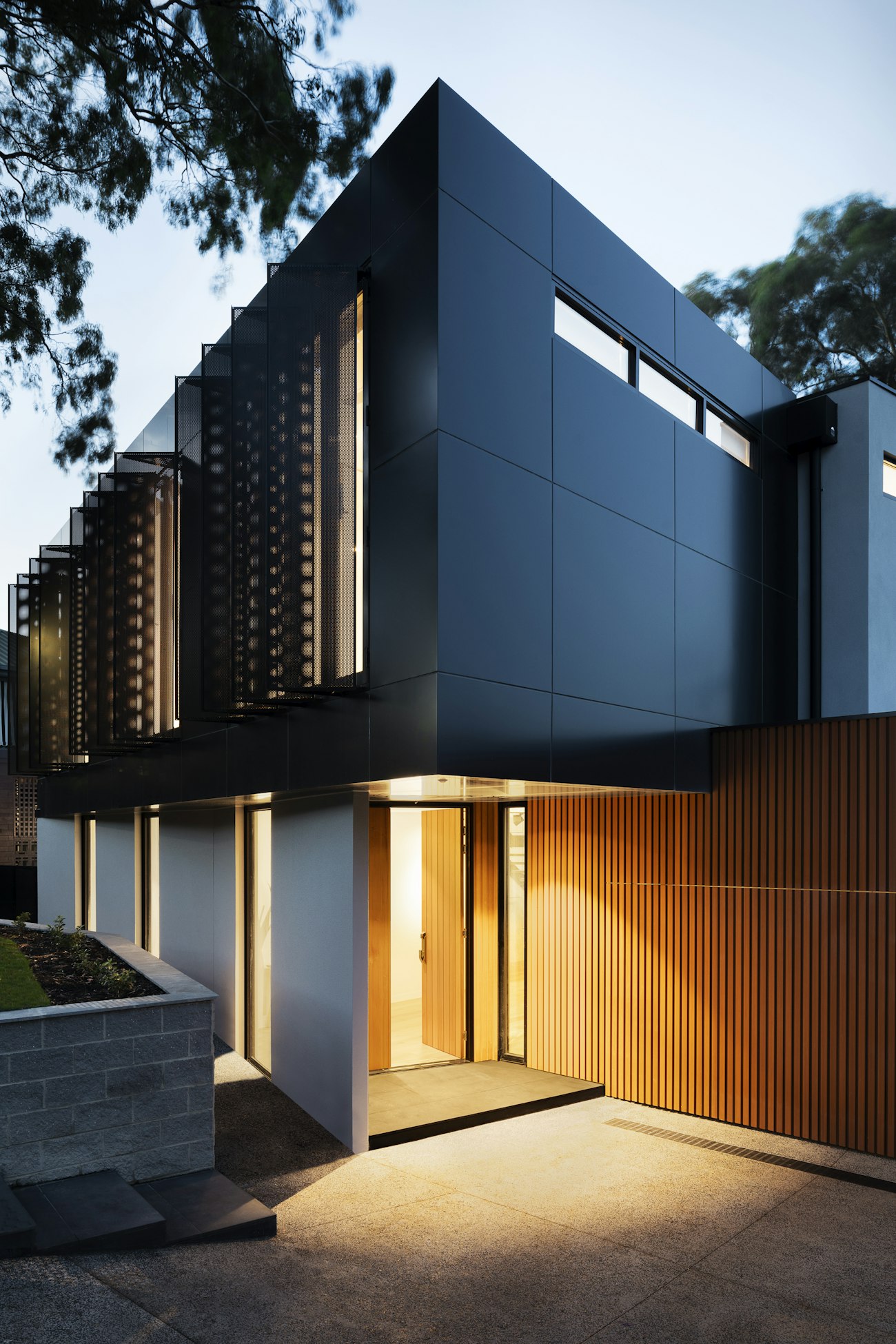 Contemporary two-storey home with black panel cladding, perforated screens and a warmly lit timber-slatted entrance at dusk