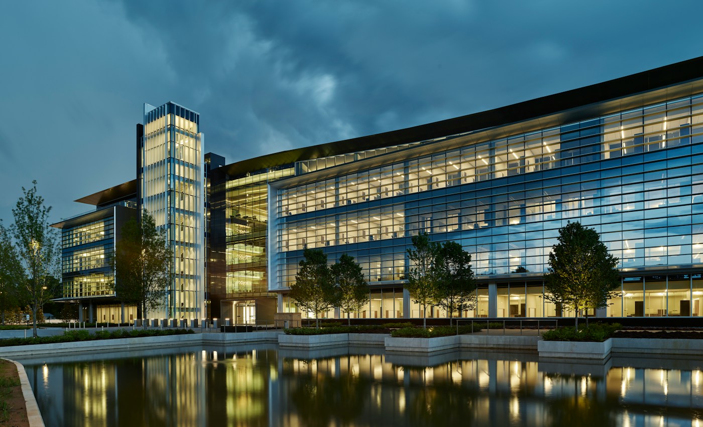 Modern glass-fronted commercial building glowing at dusk, with lit trees and a reflecting pool