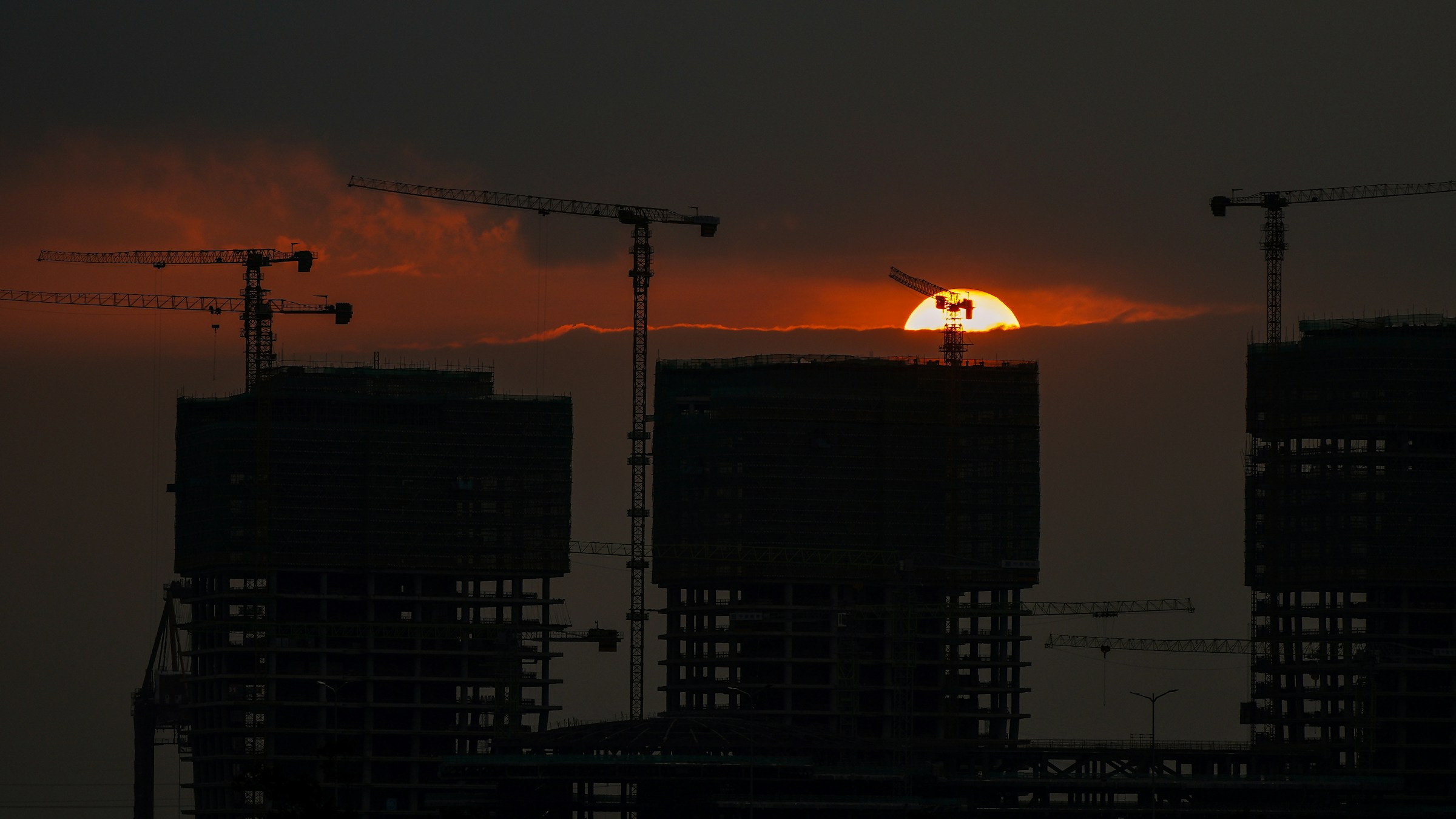 Buildings under construction with tower cranes silhouetted against a setting sun