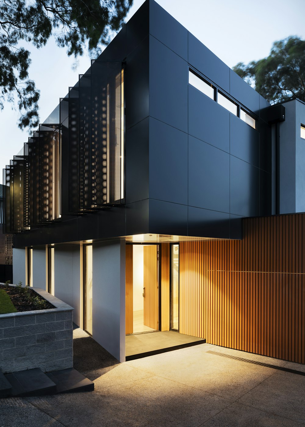 Contemporary two-storey Nexora home with a dark panelled facade, timber cladding and a warmly lit entrance at dusk