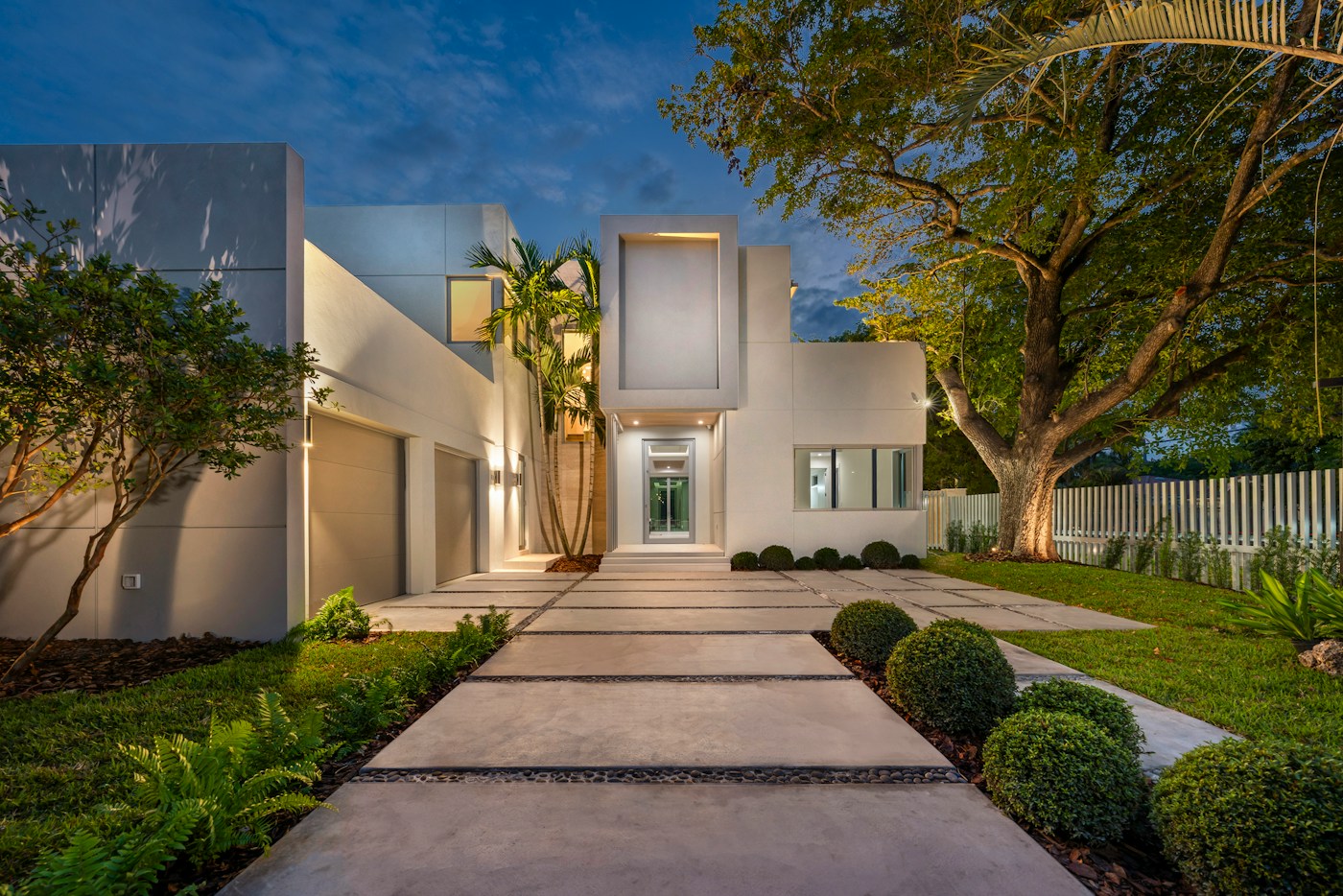 Modern Nexora-built residence with concrete facades, warm interior lighting and mature landscaping at dusk