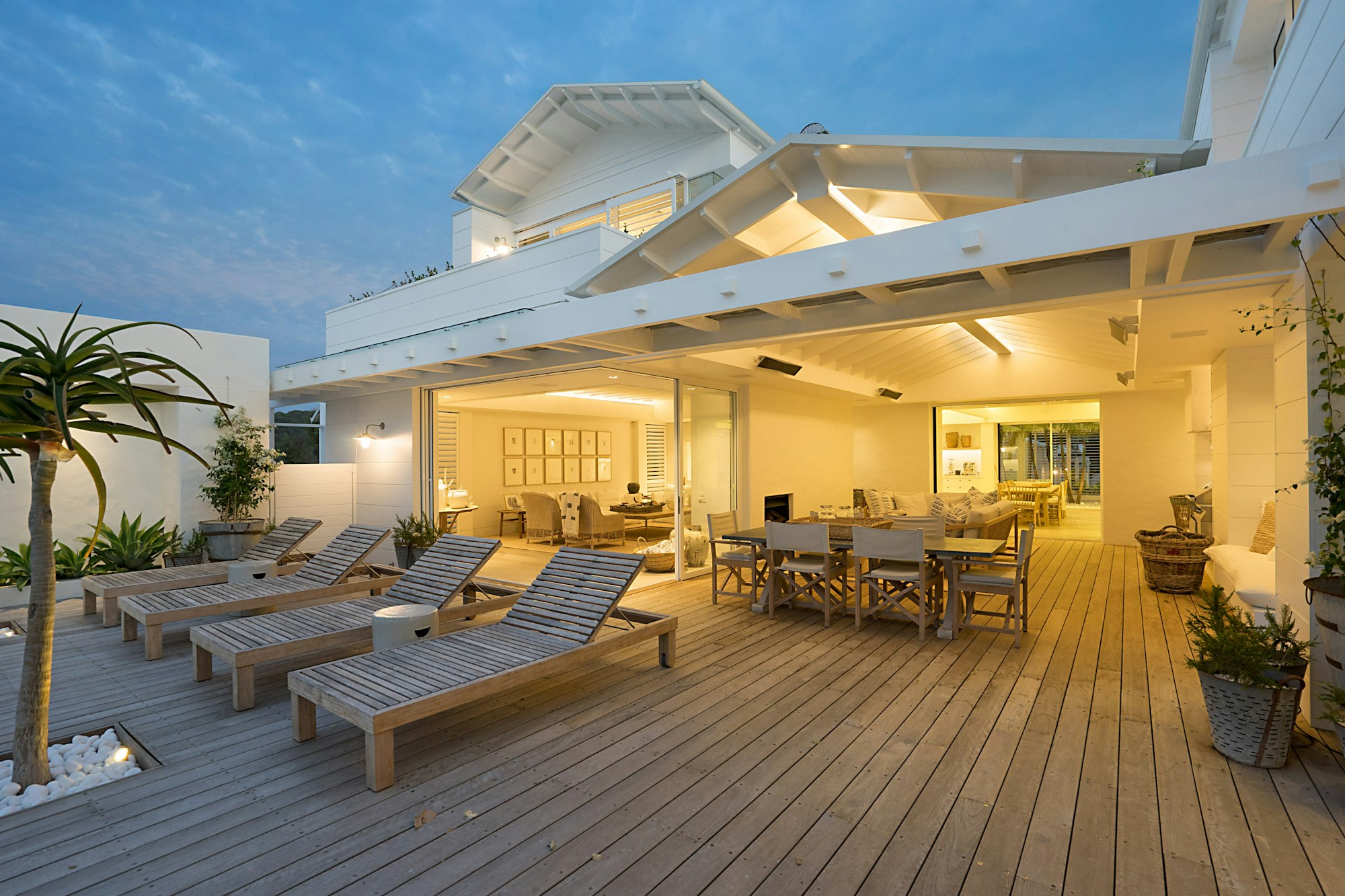 Contemporary two-storey villa at dusk with warmly lit interiors, a covered timber deck and loungers